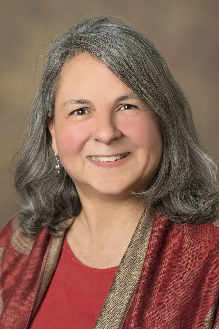 Woman with long gray hair, smiling, wearing a red and tan silky top