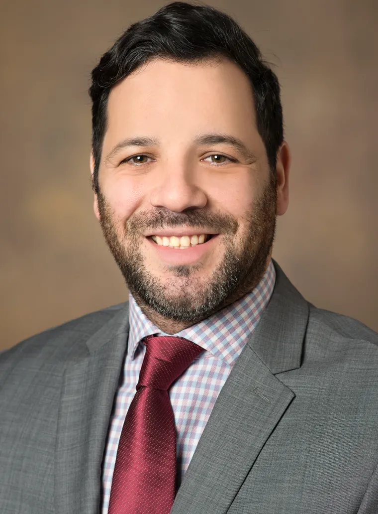 Man with short dark brown hair, smiling, wearing gray jacket, blue and white checkered shirt, and burgundy red tie